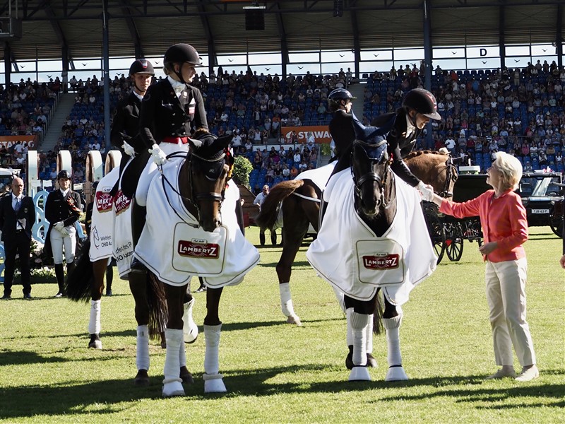 Præmieuddelingen i holdkonkurrencen fandt sted inde på det store stadion i modlys og på lånte heste! Her er det Ursula von der Lien, formanden for EU-kommissionen, som selv er en passioneret rytter, der lykønsker ankermanden Cathrine Dufour med den flotte sejr.